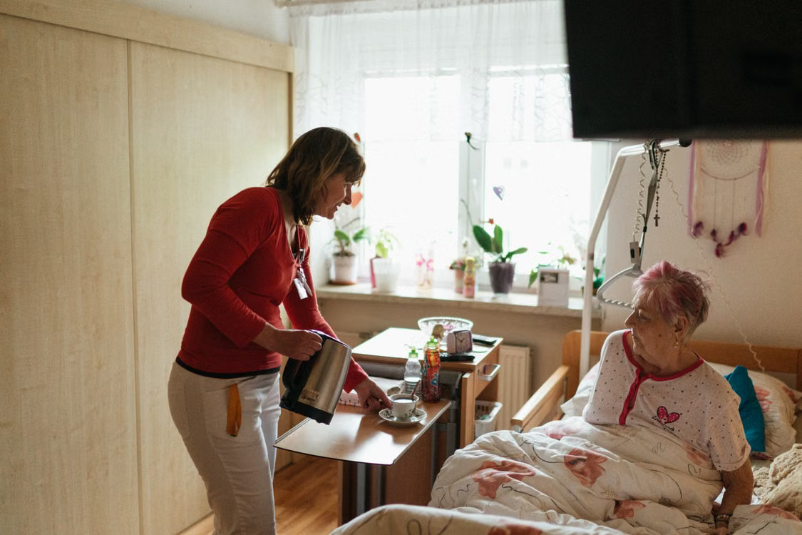 A home care worker brings a cup of tea to an elderly client resting in bed.